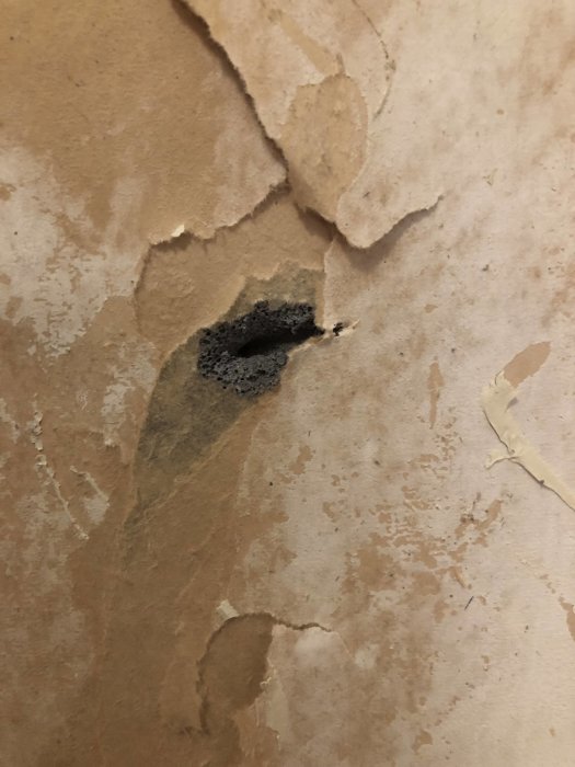 Närbild på en skadad vägg med flagnande lager som avslöjar underliggande svart material, troligtvis inte gips.