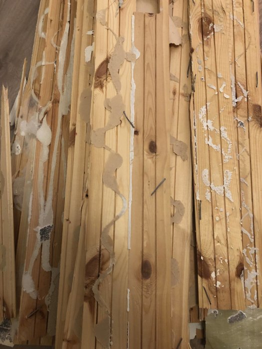 Träpanelvägg med rester av avskalat material och synliga kvistar, indikerar ej gips utan tunt skikt innan betong.