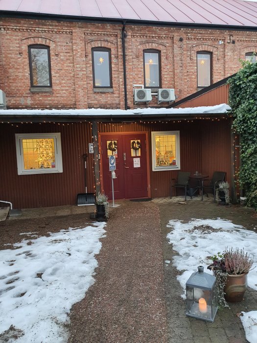 Gångväg leder till röd husdörr på tegelbyggnad med julstjärnor i fönster, belysning, snöfläckar och utomhusdekoration.