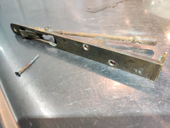 Trasig mässingsdel från ytterdörr med skruv bredvid på metallunderlag.