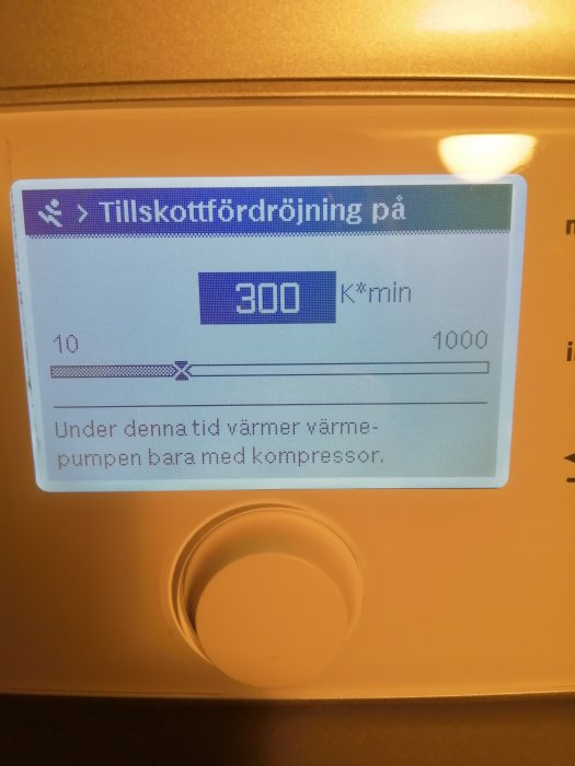 Digital display på en värmepump visar inställningen "Tillskottfördröjning på 300 K*min" och meddelandet "Under denna tid värmer värmepumpen bara med kompressor.