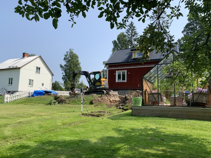 Grävmaskin gräver vid en röd hus med vit källare på en solig dag för ett renoveringsprojekt.