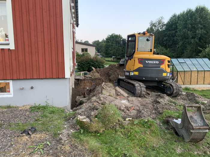 Grävmaskin utför dräneringsarbete bredvid rödmålad husvägg, jordhögar och uppriven mark syns.