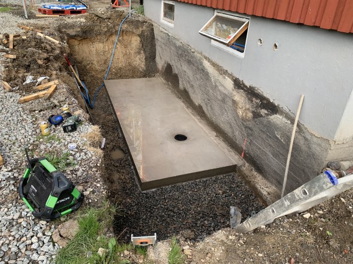 Nyanlagd källaringång med betongtrappa vid husvägg under konstruktion, omgiven av grus och byggmaterial.