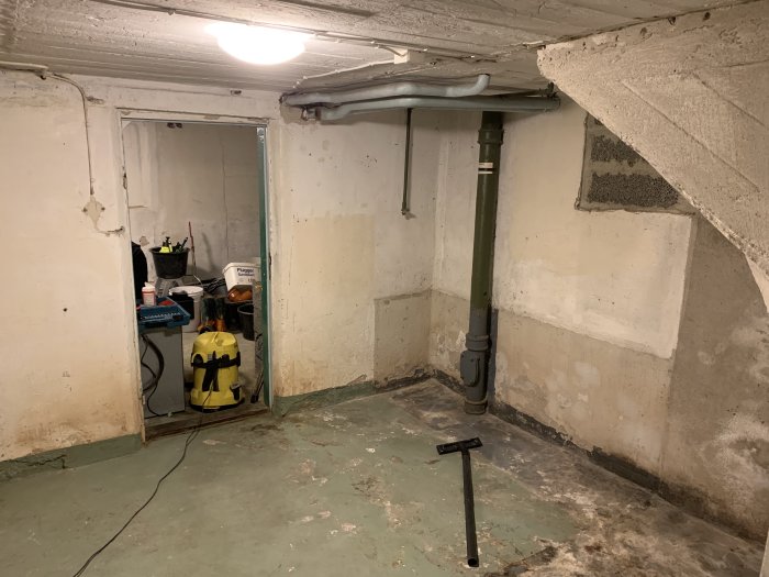 Källarrum under renovering med avloppsrör och verktyg, ommålade väggar och grovbetonggolv.