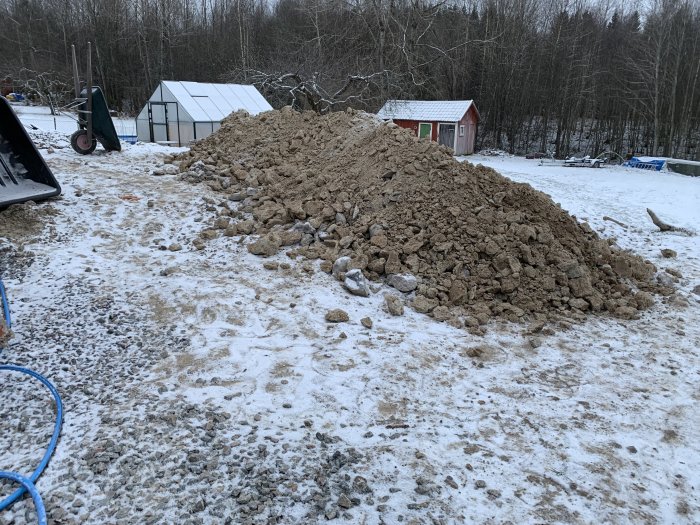 Stor hög med uppschaktad lera och grus på vintermark bredvid en skottkärra och glashus.