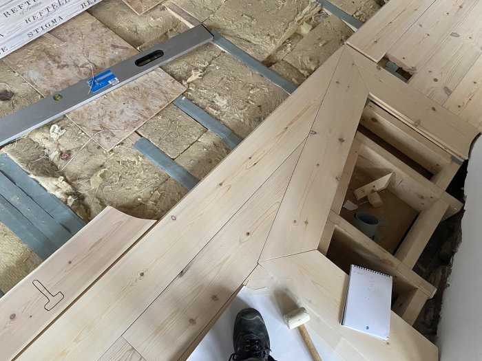 Trägolv under installation med synliga isoleringsmaterial, verktyg och anteckningsblock.