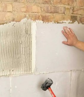 En hand trycker mot ett nyinstallerat isoleringsblock på en vägg ovanför en gummihammare, med äldre tegelsten och puts i bakgrunden.