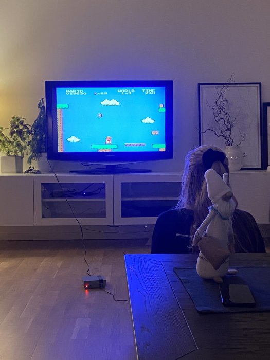 Person i vardagsrum spelar klassiskt plattformsspel på TV, med tomtefigur och konsol i förgrunden.