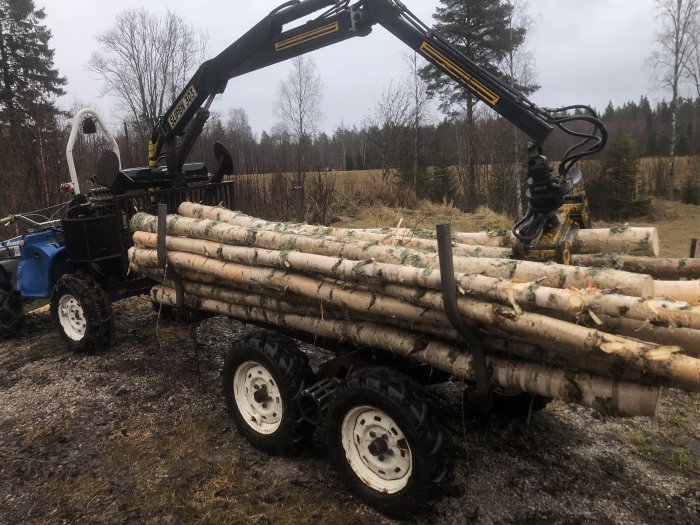 Traktor med en lastad timmervagn och kran i ett skogsområde, vilket visar utrustning för skogsbruk.