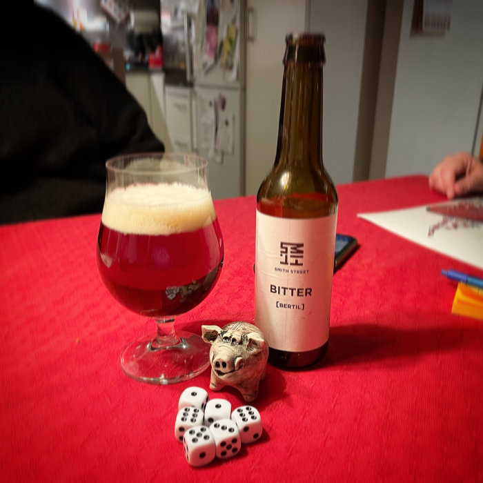 Ölglas med mörk öl, flaska med etikett 'BITTER', tärning och lerfigur av gris på röd duk.