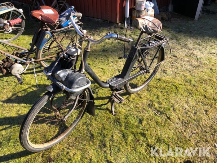 Gammal moped parkerad på gräs med synbart slitage och rost, i bakgrunden cyklar och uthus.