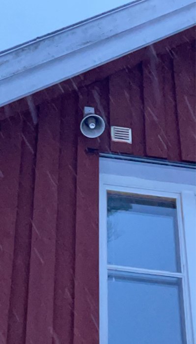 Förundrat objekt på rödmålad husvägg, ovanför ett fönster; en högtalare och en ventilkåpa.