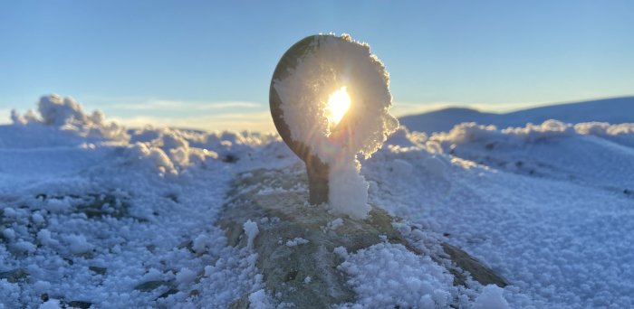 Solnedgång bakom en snöig plats med en frosttäckt metallstolpe i förgrunden.