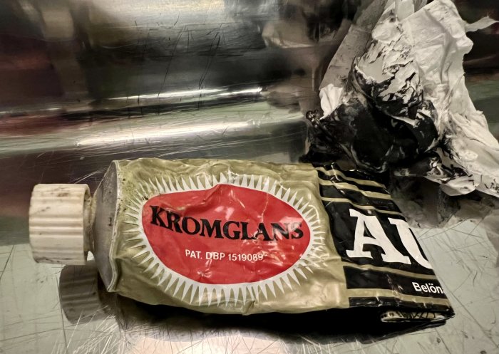 Sammansatt tub med texten 'KROMGLANS' och slitna märkningar på en repad metallisk yta.