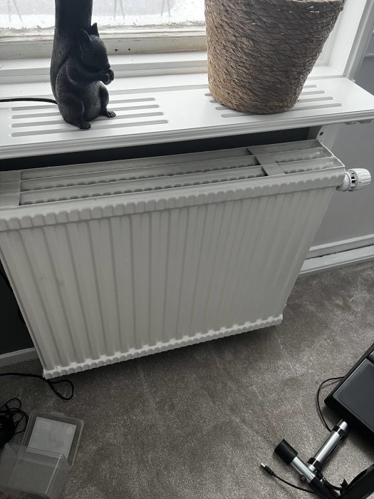 Vit radiator med termostat under ett fönster, en svart skulptur och en växt ovanpå, diverse kablar på golvet.