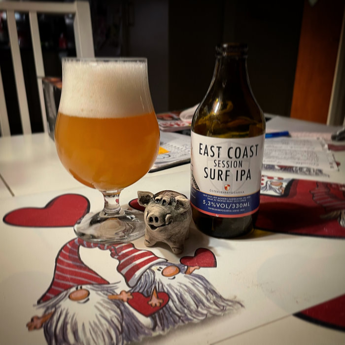 Ölglas och flaska av East Coast Session Surf IPA på bord med julduksillustrationer.