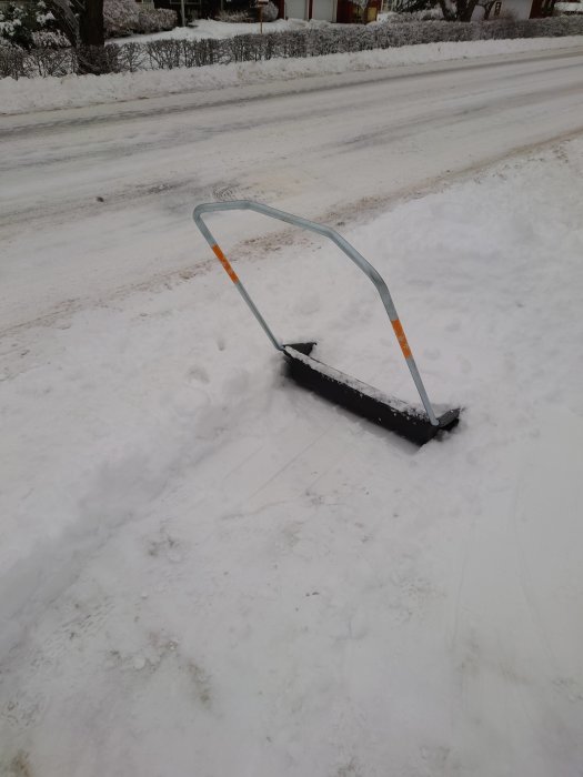En snöskyffel halvt nedsänkt i snön vid en plogad väg.