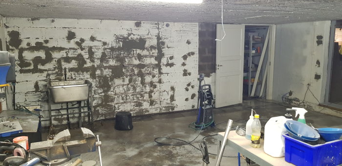 Ett garage under renovering med en tvättad och fläckig betongvägg, verktyg och rengöringsmedel på ett arbetsbord, samt en högtryckstvätt.