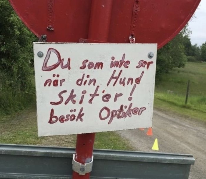Skylt med texten "Du som inte ser när din Hund skiter! besök Optiker" uppmonterad på rött föremål.