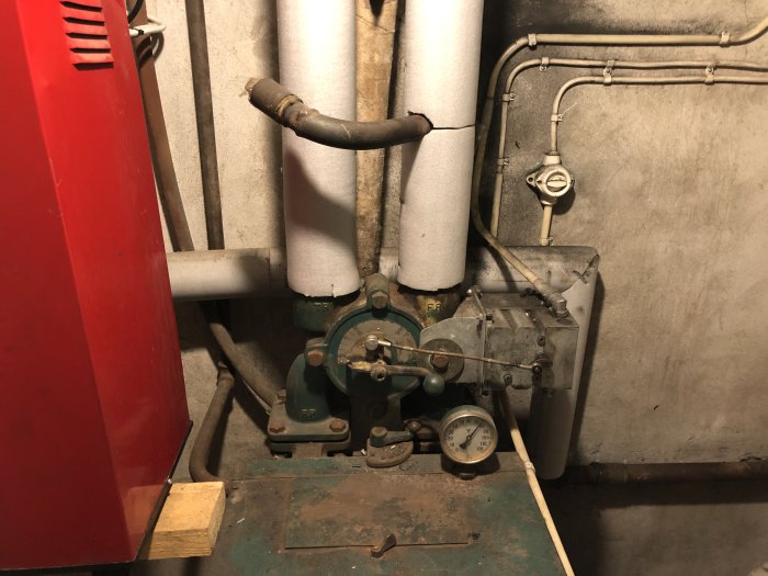 Bild på en äldre vedpanna i ett källarrum med synliga rör, manuellt reglerade ventiler och mätinstrument.