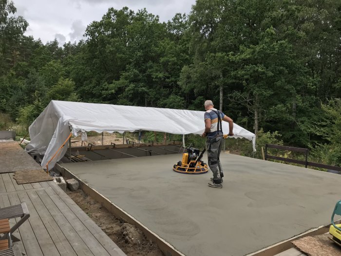 Person som slipar nygjuten betongyta under vit presenning med omgivande skog.