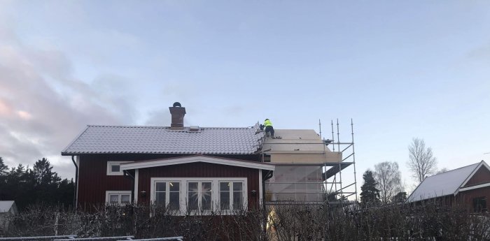 Trähus med rött tegeltak under renovering, skyddsplast och byggnadsställning syns, arbetare på taket.