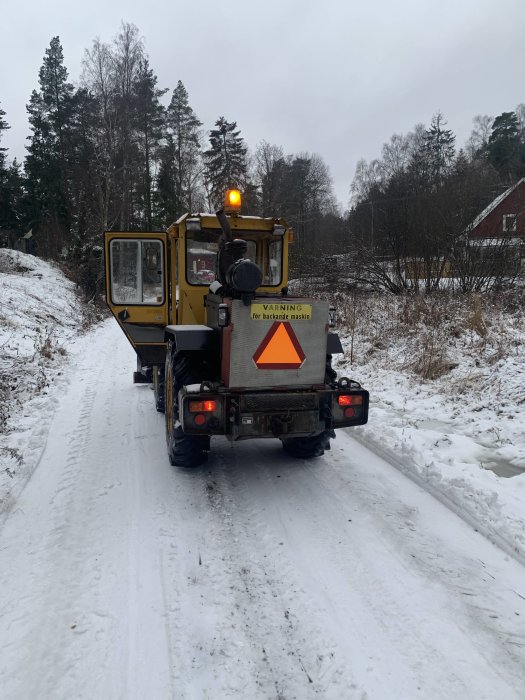 Gul vägbyggnadsmaskin i arbete på en snöig väg med skylt som varnar för backande maskin.