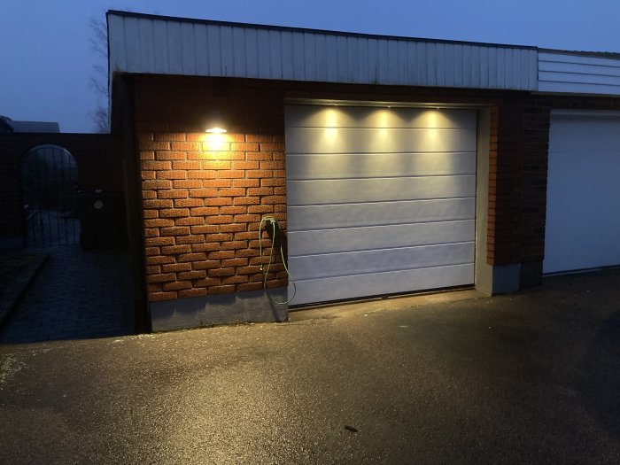 Garagefasad i skymning med en tänd vägglampa och ett stängt garageport; en bilkupévärmare är ansluten till vägguttag.