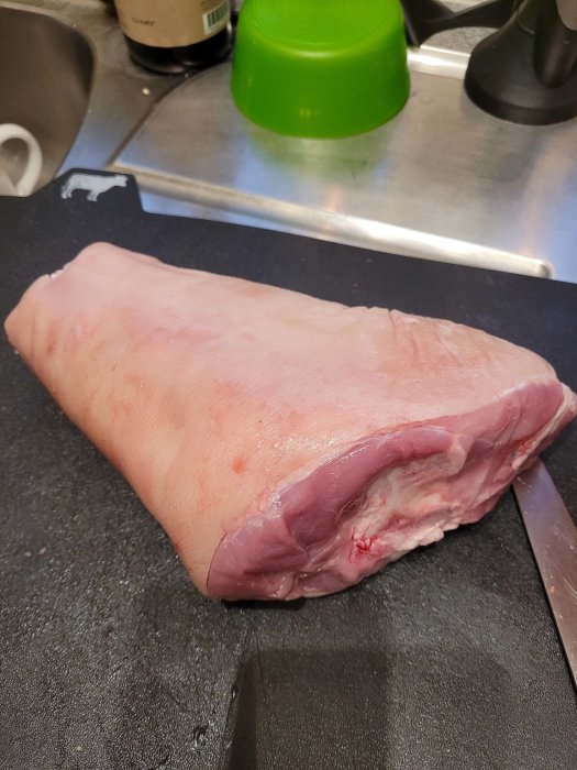 Rått stort stycke kött på en svart skärbräda bredvid en kniv och kryddburkar.