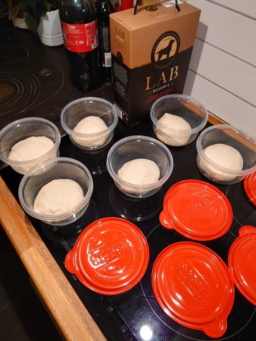 Pizzadeg i plastskålar med röda lock på ett köksbord, bredvid en vinkartong och Coca-Cola-flaska.