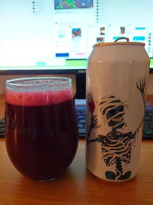 Ett glas med rödfärgad dryck och en ölburk med skelettdesign framför en datorskärm.