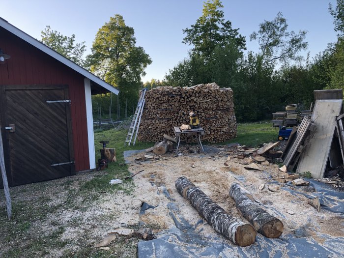 Rund vedstack bredvid röd lada, med vedkapningsplats och två stammar i förgrunden.