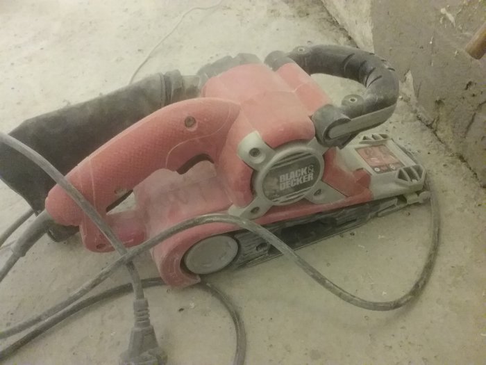 Rosa och svart bandslipmaskin av märket Black & Decker täckt med sågspån på en betongyta.