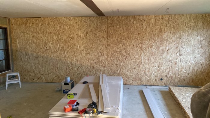 Ett rum under renovering med nyinstallerade OSB-skivor på väggen och byggmaterial på golvet.