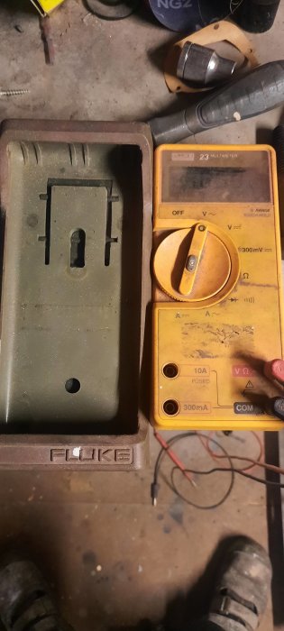 Gammal Fluke 23 multimeter med svårläst skärm, placerad i sin fodral vid en arbetsbänk, omgiven av verktyg.