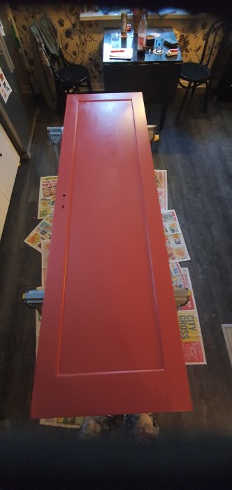 Röd målad dörr liggande på tidningspapper för torkning i ett rum med tapetserade väggar.