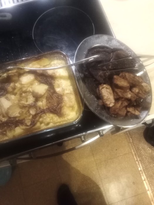 En ugnsform med potatisgratäng och en skål med stekt kött bredvid en spis på ett köksgolv.