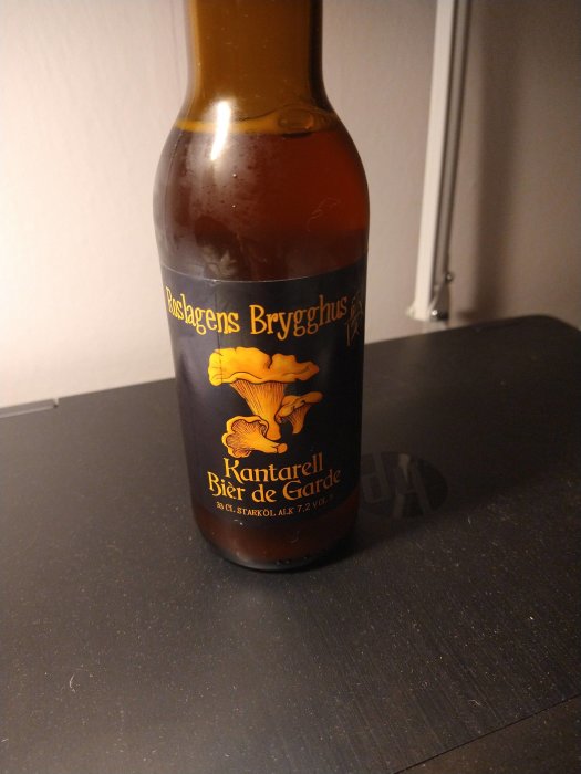 Ölflaska på bord med etikett "Roslagens Brygghus Kantarell Bier de Garde" mot en suddig bakgrund.