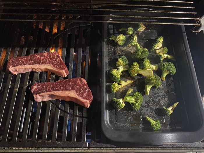 Två råa biffar på en grill bredvid en bakplåt med broccolibuketter redo att grillas.