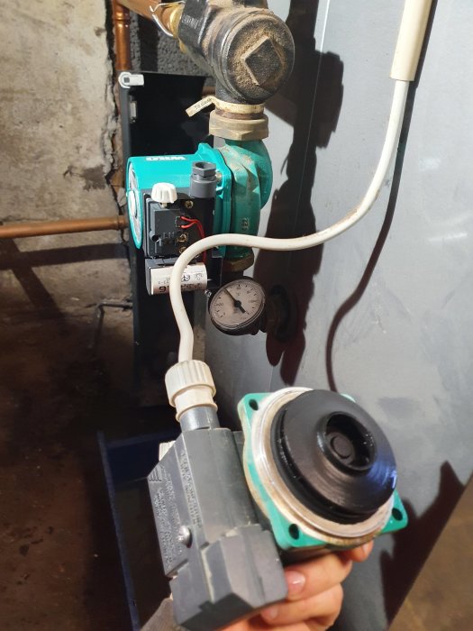 Hand som håller en demonterad pumpmotor framför en cirkulationspump och rörkopplingar med manometer.