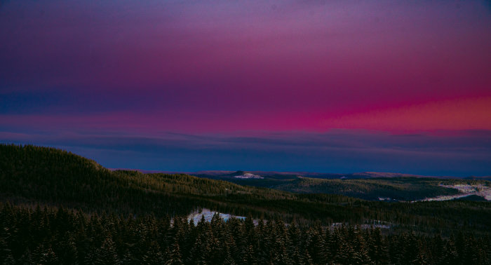 Skogklädda kullar under en dramatisk himmel med lila och rosa nyanser vid skymning.