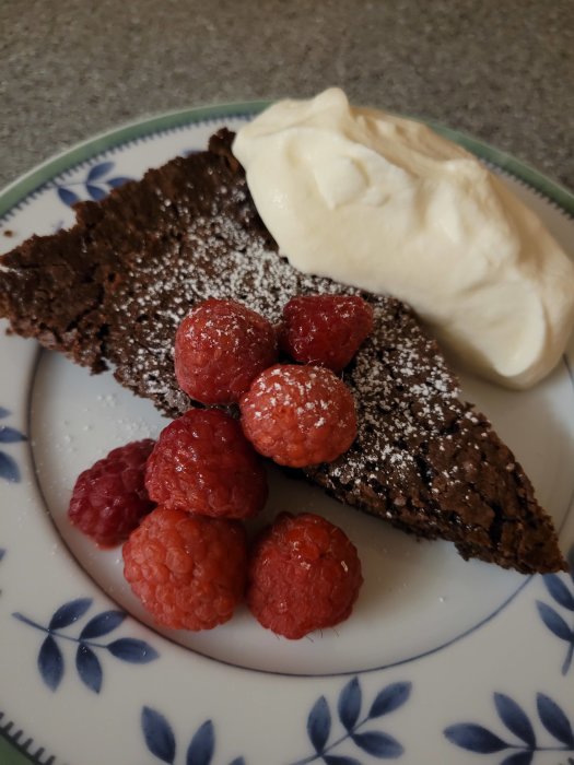 Bit av kladdkaka med pudrat florsocker, färska hallon och en klick grädde på en mönstrad tallrik.