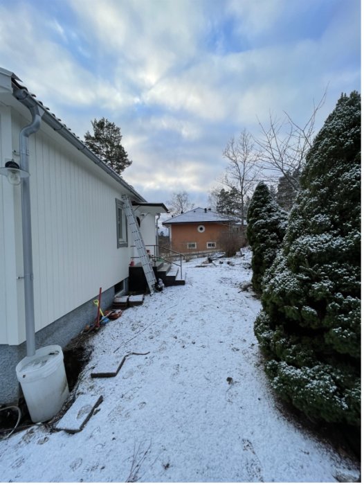 Baksidan av ett hus med lätt snötäcke, renoveringsmaterial vid gaveln och en buskrad i förgrunden.
