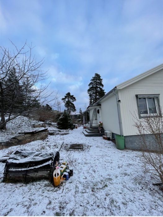 Snöbelagd trädgård vid ett hus med berg i bakgrunden och en grävskopa framför, potentiell dräneringsplats.
