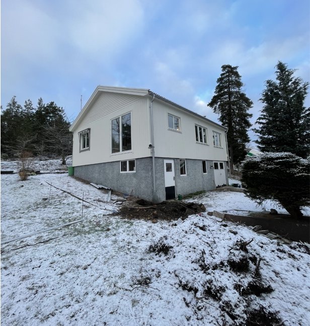 Ett ljusfärgat sutteränghus med synligt berg bakom och spår efter markarbete i snötäckt trädgård.