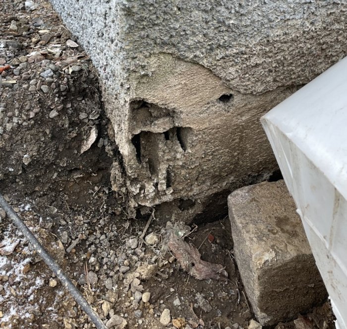 Eroderad betonghålsten vid husgrund med synlig jordskada och grus, vilket indikerar tidigare markarbeten.