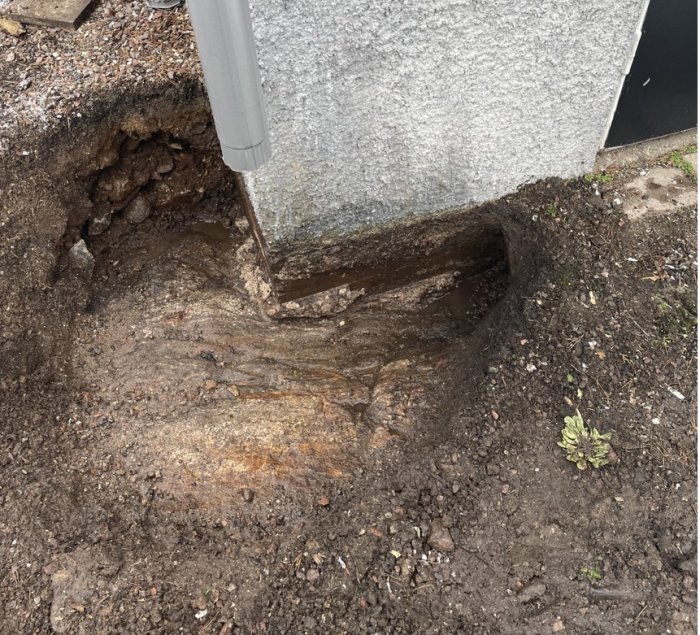 Uppgrävd jord vid husgrund som visar eroderad betonghålsten och berggrund.
