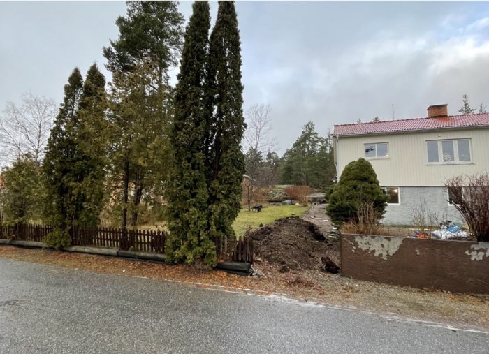 Utsikt över en tomt med påbörjade grävarbeten nära en vit husfasad, omgiven av gröna träd och en brun stenmur längs vägen.