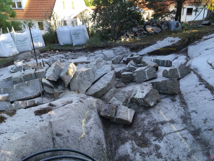 Kuperad tomt med granitklippor och sprängda stenblock, gulmarkerad för planering av byggprojekt, med hus och trädgård i bakgrunden.